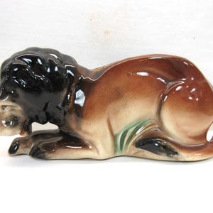 May include: A ceramic figurine of a brown and black lion lying down with its head resting on its paws. The lion has a green and brown glaze on its body.