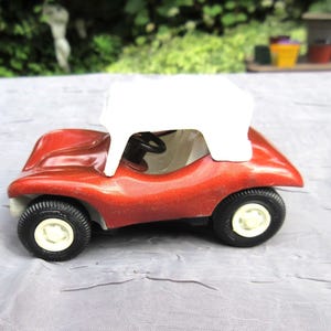 Puede incluir: Un coche de juguete rojo vintage con techo blanco. El coche tiene neumáticos negros con llantas blancas y un volante negro. Un diseño de juguete clásico.