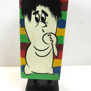 May include: A vintage wooden figurine of a cartoon character with a white body, black hair, and a worried expression. The figure is on a black base, with a colorful striped background of red, green, blue, and yellow.
