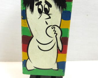 May include: A vintage wooden figurine of a cartoon character with a white body, black hair, and a worried expression. The figure is on a black base, with a colorful striped background of red, green, blue, and yellow.