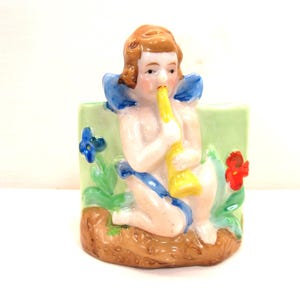 May include: A ceramic wall planter featuring a figurine of a cherub playing a yellow trumpet. The cherub is wearing a blue sash and is surrounded by green leaves and blue and red flowers.