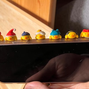 Puede incluir: Una colección de figuras de pollitos amarillos, cada una con un sombrero diferente, dispuestas sobre un teléfono inteligente. Los sombreros incluyen un gorro de Papá Noel, una gorra azul, un sombrero blanco y amarillo, un sombrero de rana azul, un sombrero con temática de abeja y un lazo rojo.