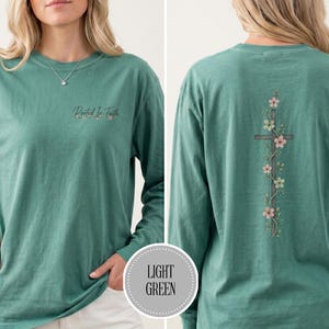 May include: A long-sleeve teal shirt with a crew neckline. The front features the text "Rooted In Faith" in small cursive script. The back displays a vertical wooden cross graphic entwined with delicate pink and white flowers and green leaves.