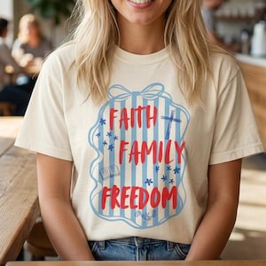 Puede incluir: Camiseta color crema con la frase "Faith Family Freedom" en rojo y azul. El diseño incluye una cruz, estrellas y rayas. La persona lleva vaqueros azules y está sentada en una mesa de madera.