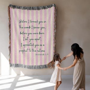 Pode incluir: Uma manta tecida listrada rosa e branca com o texto "Before I formed you in the womb I knew you...Jeremiah 1:5". A manta tem uma borda com franjas e está pendurada em uma parede.