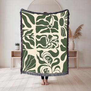 May include: A cream-colored throw blanket with a repeating pattern of green floral and abstract shapes. The blanket has a dark green fringe border. The design features stylized flowers, leaves, and other organic forms. The blanket is displayed indoors.
