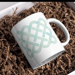 May include: White ceramic mug with a light blue Celtic knot design. The mug is inside a white box filled with brown crinkle paper, ideal for gifting. The mug has a curved handle.