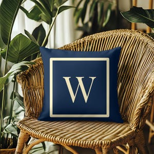 Op de afbeelding: Een marineblauw vierkant sierkussen met een crèmekleurige rand en een grote crèmekleurige letter "W" in het midden. Het kussen ligt op een rotan stoel, met groene planten op de achtergrond.