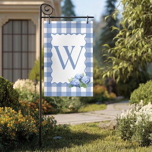May include: A garden flag with a blue and white gingham pattern background. The flag features a white scalloped frame with a large blue letter "W" and a cluster of blue hydrangea flowers. The flag is displayed on a black metal stand.