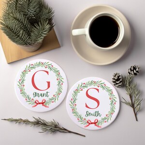 May include: Two round white ceramic coasters featuring a green holiday wreath with red berries and a red bow. One coaster displays the letter G and the name Grant, while the other shows the letter S and the name Smith in a red and green font.