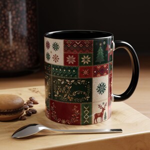 May include: A ceramic mug with a black interior and handle, featuring a festive patchwork design of red, green, and cream squares. The pattern includes snowflakes, holly, a reindeer, and a Dala horse. A silver spoon lies in front of the mug.