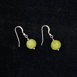 May include: A pair of earrings with silver hooks and round, light green beads. The earrings are displayed against a black background, highlighting the jewelry's simple design and color.