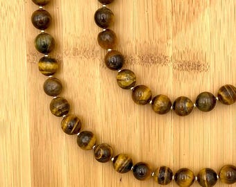 May include: Two strands of a beaded necklace with tiger eye stones. The beads are round, with a mix of brown, gold, and black colors. The necklace is displayed on a light brown wooden surface.