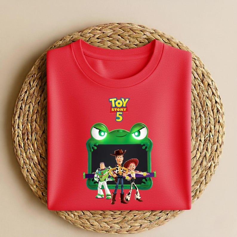 May include: A red crew neck t-shirt featuring a graphic of Toy Story characters. The design includes Buzz Lightyear, Woody, and Jessie, framed by a green frog with the text "Toy Story 5". The shirt is folded on a woven mat.