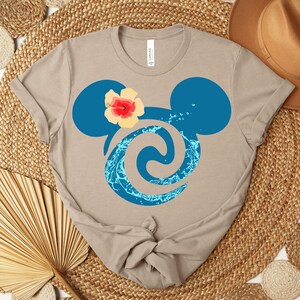 Puede incluir: Una camiseta color beige con un dise&ntilde;o azul de la cabeza de Mickey Mouse y un detalle floral. El dise&ntilde;o incorpora un remolino acu&aacute;tico. La camiseta est&aacute; anudada en la parte inferior y sobre una estera tejida.