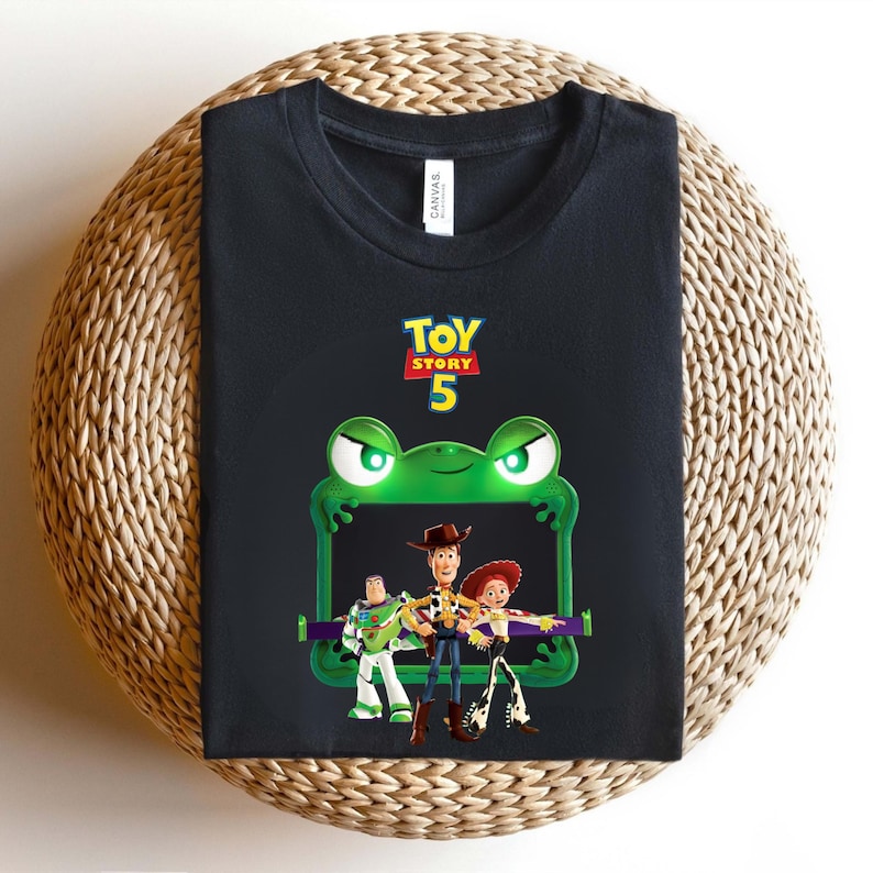 May include: Black t-shirt featuring a green frog-shaped frame with the text "TOY STORY 5". Inside the frame are cartoon characters Woody, Buzz Lightyear, and Jessie. The shirt is displayed on a woven surface.