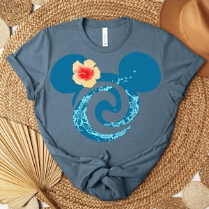 Puede incluir: Camiseta azul con un dise&ntilde;o estilizado de orejas de Mickey, un remolino de agua y una flor de hibisco. El dise&ntilde;o es en tonos azules y amarillos. La camiseta es de material suave.
