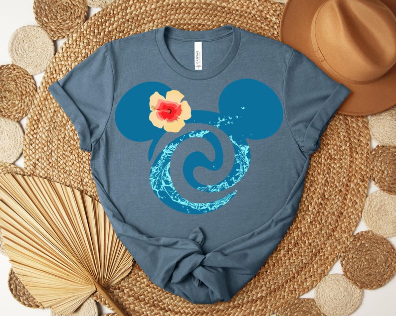 Puede incluir: Camiseta azul con un dise&ntilde;o estilizado de orejas de Mickey, un remolino de agua y una flor de hibisco. El dise&ntilde;o es en tonos azules y amarillos. La camiseta es de material suave.