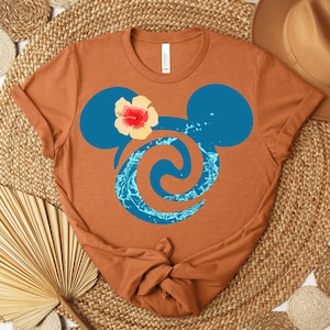 Puede incluir: Una camiseta de color &oacute;xido con un dise&ntilde;o de cabeza de Mickey Mouse azul, un remolino de agua y una flor de hibisco. La camiseta est&aacute; anudada en la parte inferior. El dise&ntilde;o est&aacute; inspirado en una pel&iacute;cula de animaci&oacute;n popular.