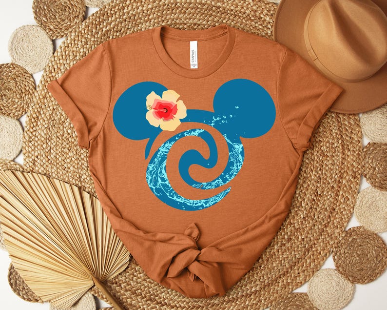 Puede incluir: Una camiseta de color &oacute;xido con un dise&ntilde;o de cabeza de Mickey Mouse azul, un remolino de agua y una flor de hibisco. La camiseta est&aacute; anudada en la parte inferior. El dise&ntilde;o est&aacute; inspirado en una pel&iacute;cula de animaci&oacute;n popular.