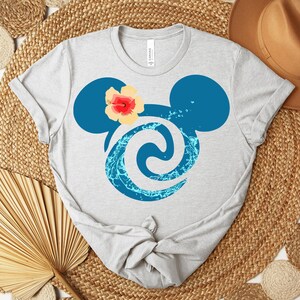 Puede incluir: Camiseta gris claro con una silueta azul de Mickey Mouse, un dise&ntilde;o de remolino de agua y una flor amarilla y roja. La camiseta est&aacute; hecha de un material suave y tiene cuello redondo.