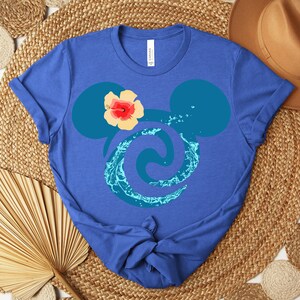 Puede incluir: Camiseta azul con un dise&ntilde;o estilizado de orejas de Mickey Mouse, un remolino de agua y una flor de hibisco. La camiseta es de material suave y est&aacute; anudada en la parte inferior.