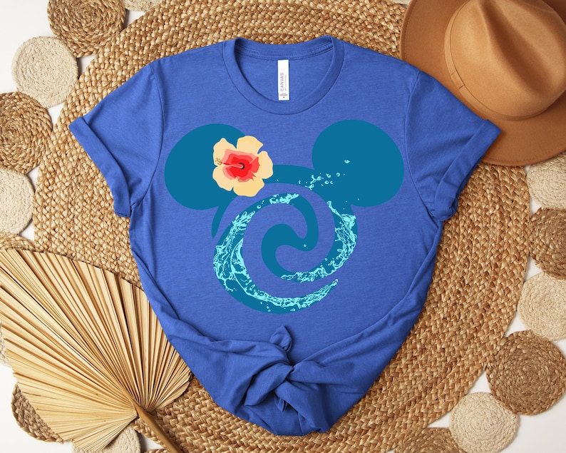 Puede incluir: Camiseta azul con un dise&ntilde;o estilizado de orejas de Mickey Mouse, un remolino de agua y una flor de hibisco. La camiseta es de material suave y est&aacute; anudada en la parte inferior.