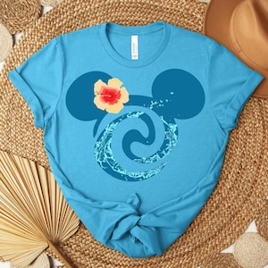 Puede incluir: Una camiseta azul claro con un dise&ntilde;o estilizado. El dise&ntilde;o incorpora orejas de Mickey Mouse, un remolino acu&aacute;tico y una flor. La camiseta est&aacute; hecha de un material suave y tiene cuello redondo.