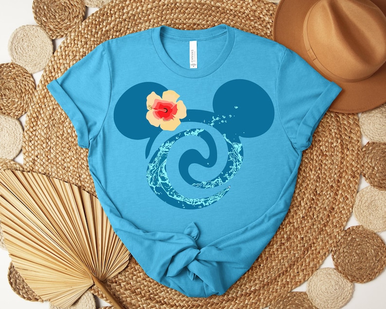 Puede incluir: Una camiseta azul claro con un dise&ntilde;o estilizado. El dise&ntilde;o incorpora orejas de Mickey Mouse, un remolino acu&aacute;tico y una flor. La camiseta est&aacute; hecha de un material suave y tiene cuello redondo.