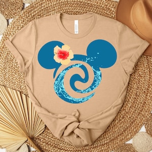 Puede incluir: Camiseta color beige con un dise&ntilde;o de cabeza de Mickey Mouse azul, una flor de hibisco y un remolino de agua. La camiseta est&aacute; anudada en la parte inferior y se muestra sobre una estera tejida con elementos decorativos.