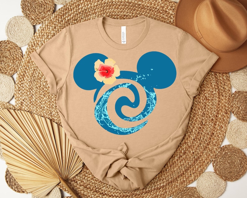 Puede incluir: Camiseta color beige con un dise&ntilde;o de cabeza de Mickey Mouse azul, una flor de hibisco y un remolino de agua. La camiseta est&aacute; anudada en la parte inferior y se muestra sobre una estera tejida con elementos decorativos.