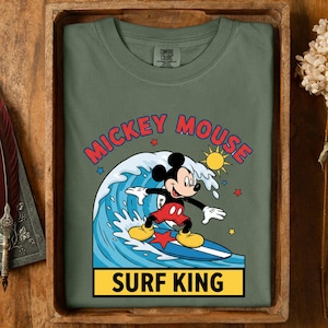 May include: Olive green t-shirt featuring a graphic of Mickey Mouse surfing a wave. The design includes the text "Mickey Mouse" in red and "Surf King" in yellow. The shirt is a Comfort Colors brand.