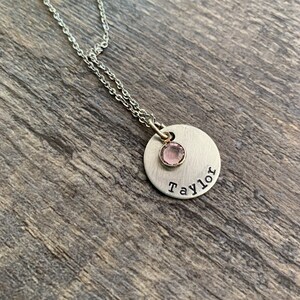 May include: A silver necklace with a round pendant that says "Taylor" and has a small pink gemstone in the center.