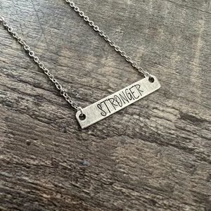 May include: A silver necklace with a silver bar pendant that says "STRONGER" on it.