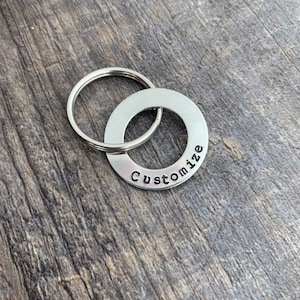 May include: A silver-colored metal keychain with a circular tag that reads "Customize" in a black, stamped font. The keychain has a split ring for attaching keys. The background is a wooden surface.