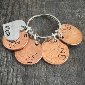 May include: A silver heart charm with the word "Mom" engraved on it, attached to a silver key ring with five copper pennies. Each penny has a heart and a letter engraved on it: K, B, I, N, and Z.