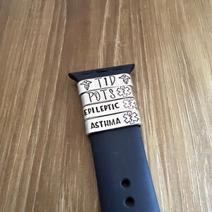 May include: A black Apple Watch band with four silver medical alert bands. The bands read "TID", "POTS", "EPILEPTIC", and "ASTHMA". Each band has a medical symbol.