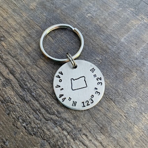 May include: Silver keychain with an outline of the state of Oregon and the coordinates 44.04° N 123.03° W engraved on a round tag.