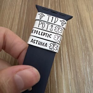 May include: A black Apple Watch band with five silver metal bands. The bands have the words "T1D", "POTS", "EPILEPTIC", "ASTHMA", and a medical symbol.