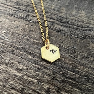 May include: A gold-colored necklace with a hexagonal pendant. The pendant features a small, black bee illustration. The necklace chain is delicate and hangs on a wooden surface.