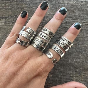 May include: A collection of silver-toned metal rings worn on fingers. The rings feature various stamped designs including a sun, mountains, boots, leaves, a compass, fish, and the names Paul, Emma, and Ruby. Each ring has an open-band design.