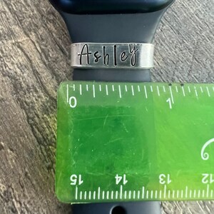 May include: A silver watch band with the name "Ashley" engraved on it. The band is shown next to a ruler, measuring approximately 2.5 cm in width.