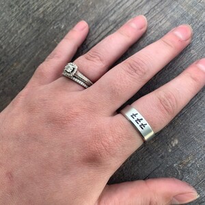 May include: A silver ring with the word "Love" engraved on it. The ring is worn on the ring finger of a person's left hand. The person is also wearing a diamond engagement ring on the same finger.