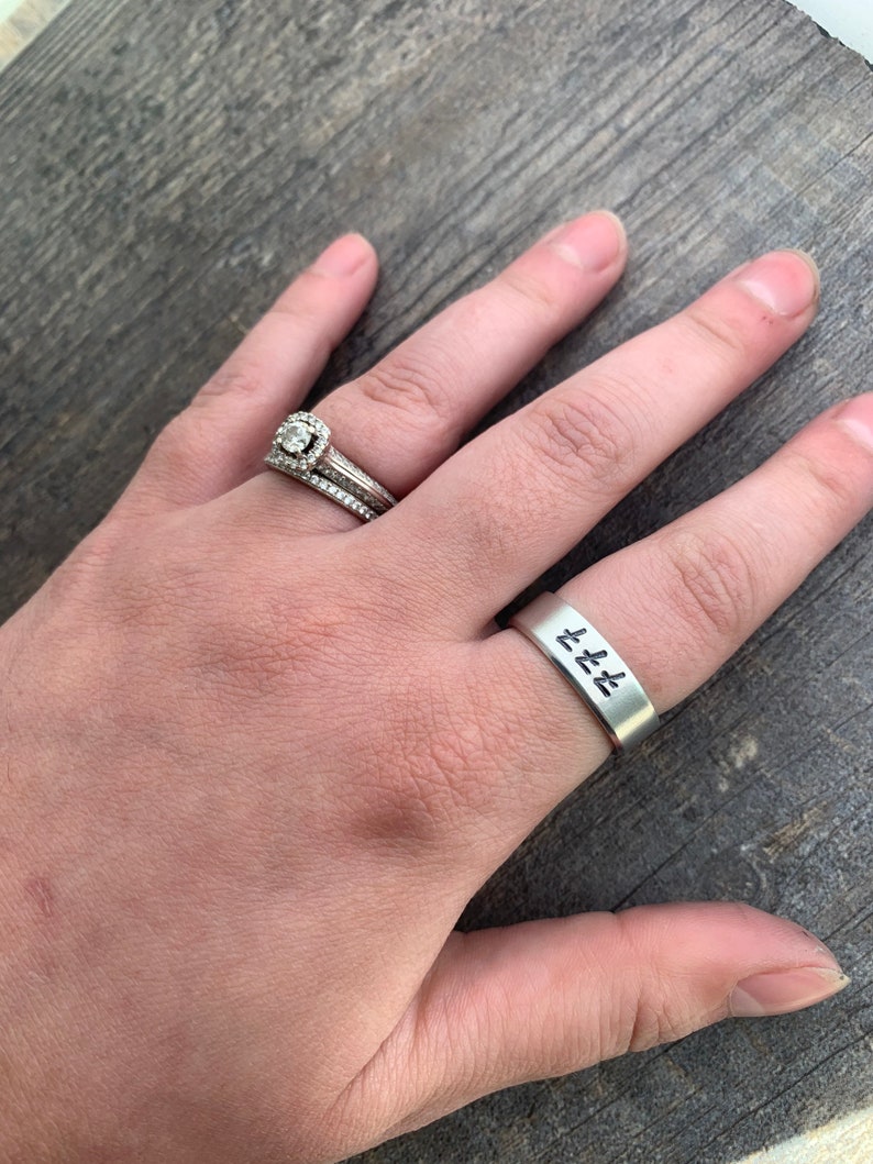 May include: A silver ring with the word "Love" engraved on it. The ring is worn on the ring finger of a person's left hand. The person is also wearing a diamond engagement ring on the same finger.