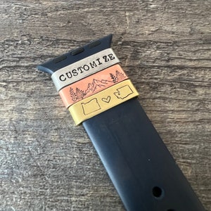 May include: A black watch band with three metal bands attached. The top band is silver and says "CUSTOMIZE". The middle band is copper with a mountain range design. The bottom band is gold with an outline of the state of Oregon and a heart.