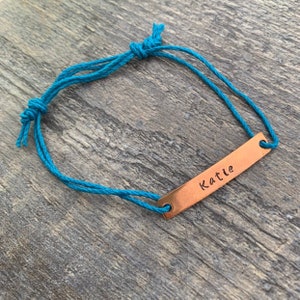 May include: A turquoise cord bracelet with a copper bar charm engraved with the name "Katie".