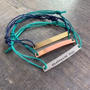 May include: Three bracelets with rectangular metal bars and adjustable cord ties. One bar is gold, one is copper, and one is silver with the text "customize this". The cords are teal and navy blue.