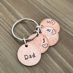 May include: A copper keychain with a silver ring. The keychain has a round tag stamped with the word "Dad" and several copper coin charms. The coins are stamped with initials and heart symbols. The background is a wooden surface.