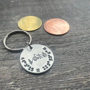 Può includere: Un portachiavi in ​​argento con un design a catena montuosa e coordinate incise. Le coordinate sono 40.315 N 123.053 W.