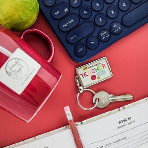 Op de afbeelding: Een rode mok, een limoen, een blauw toetsenbord, een sleutelhanger met de tekst "Best Mrs. Kanya TEACHER ever", een sleutel, een potlood en een open planner op een rode ondergrond. De planner staat open op week 40.